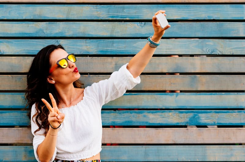 pretty woman wearing white shirt in front of multi-colored wood boards taking a selfie with smartphone camera and holding up peace symbol to help with her online presence
