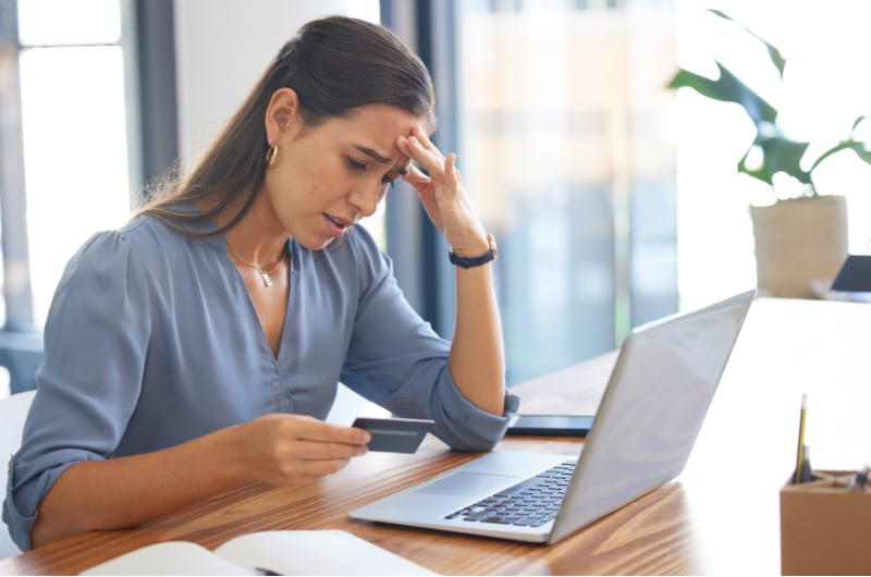 women sitting at a desk with computer and holding credit card looking stressed for financial hardship